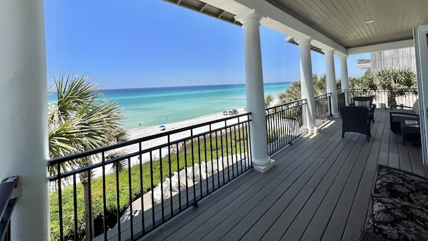 Un espace extérieur abrité, avec vue sur la plage, comprenant une terrasse en bois, des colonnes blanches et une balustrade.