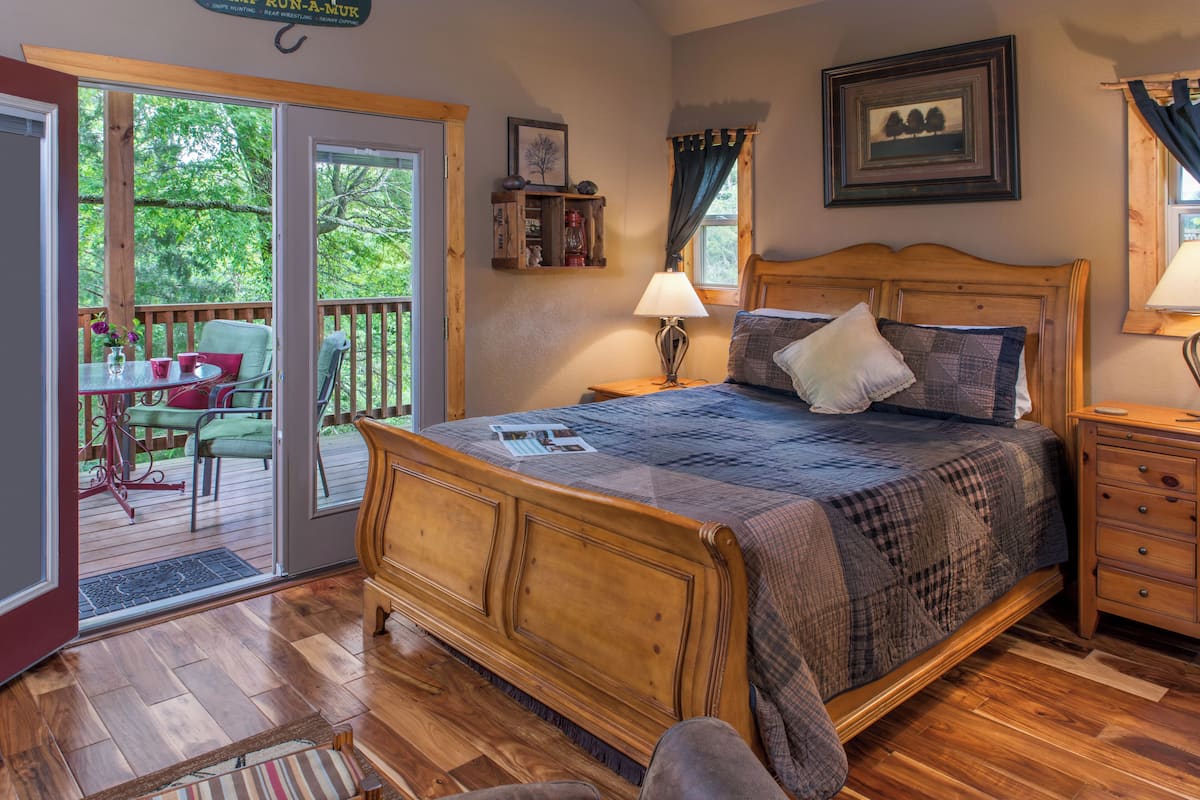 A bedroom with a wooden bed, a nightstand, a lamp, a window with curtains, a door leading to a balcony, and a small table with chairs outside.