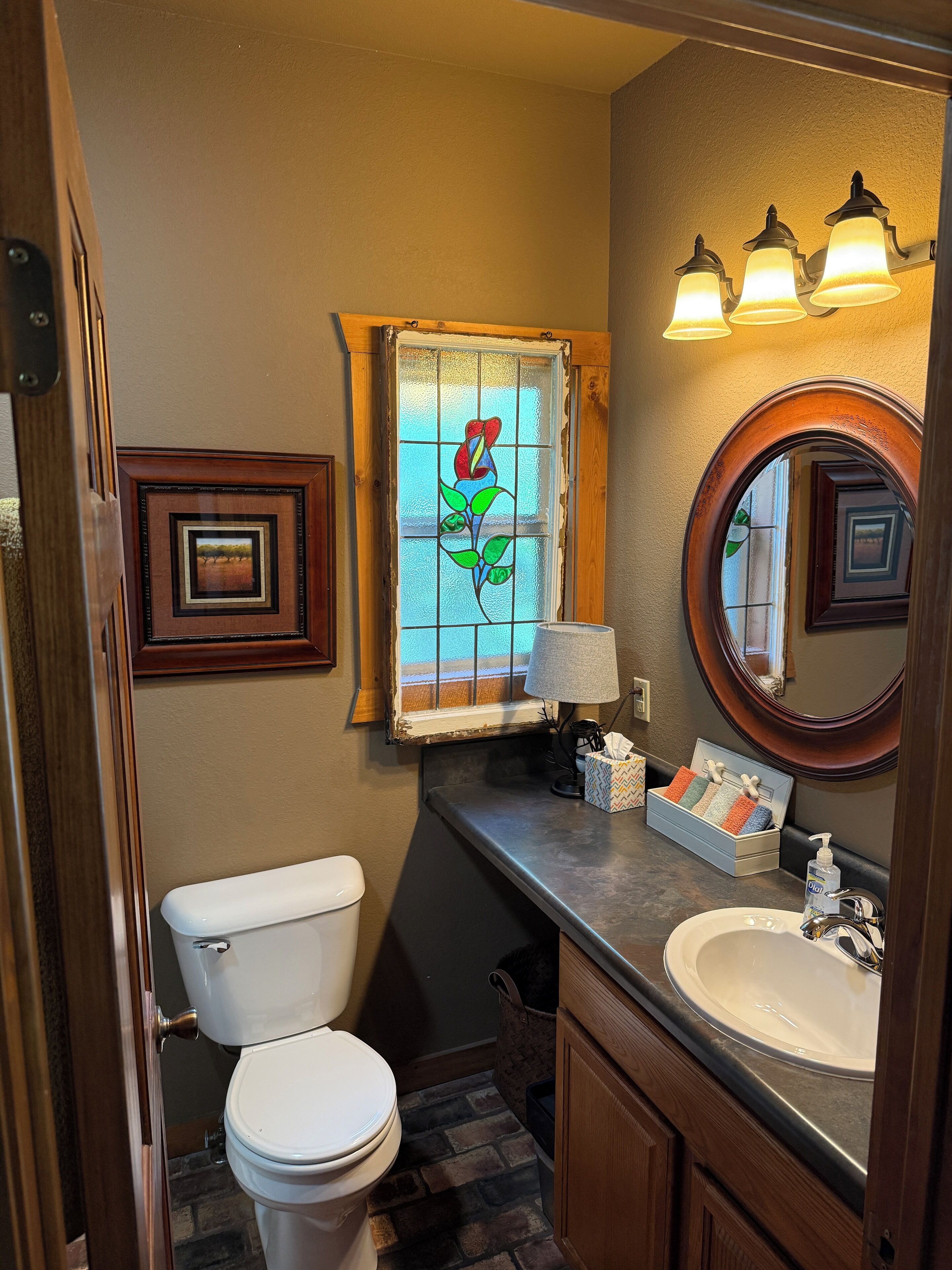 A bathroom with a white toilet, a sink with a mirror, a stained glass window, and a small framed picture on the wall.