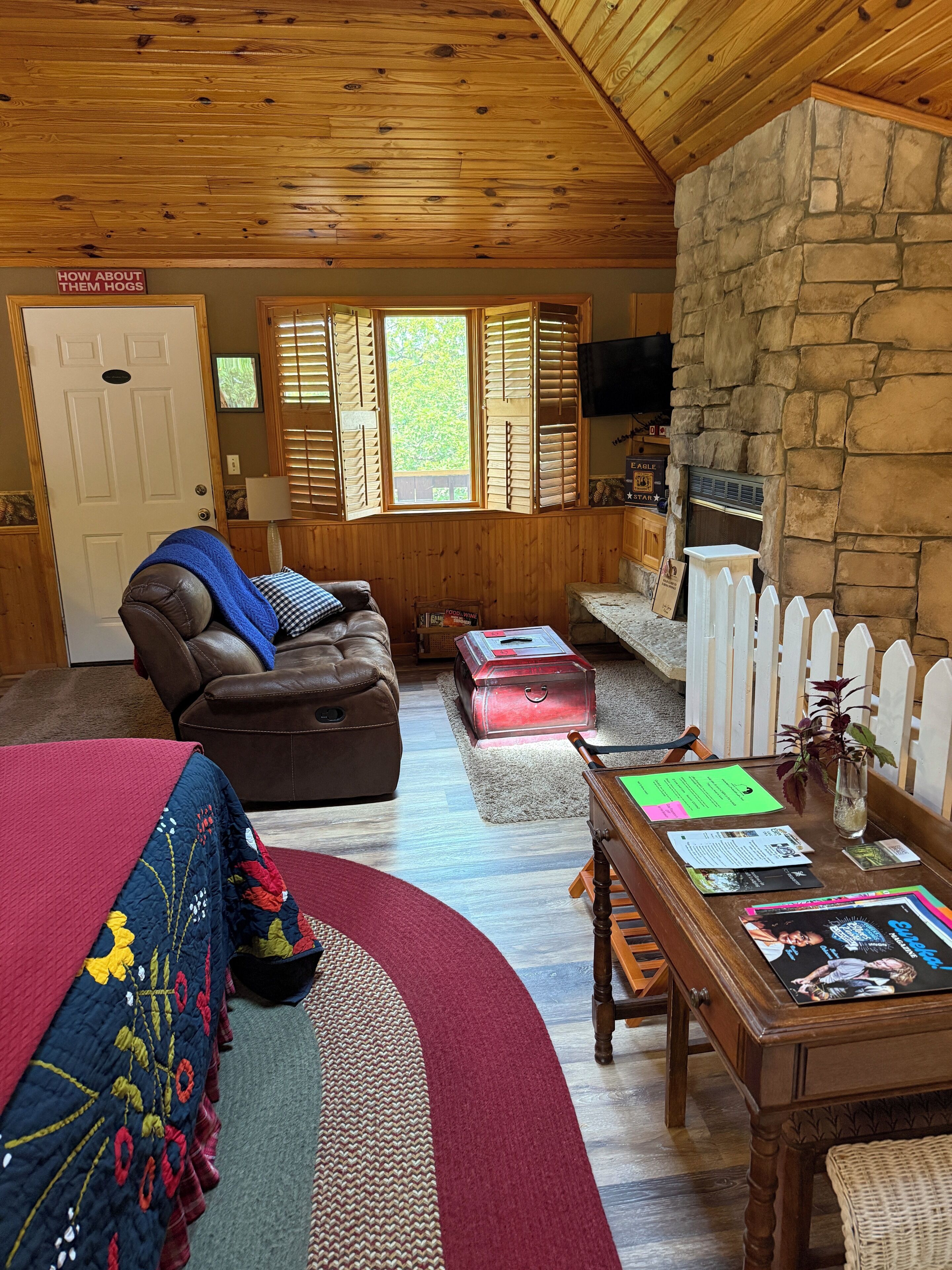 A cozy cabin interior with a leather sofa, a dining table with papers, a stone wall, and a wooden ceiling.