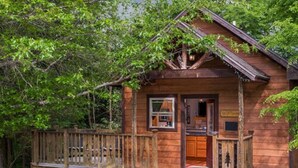 A wooden cabin with a porch and a small deck, surrounded by trees. - Eureka Sunset Cabins (Eureka Springs)