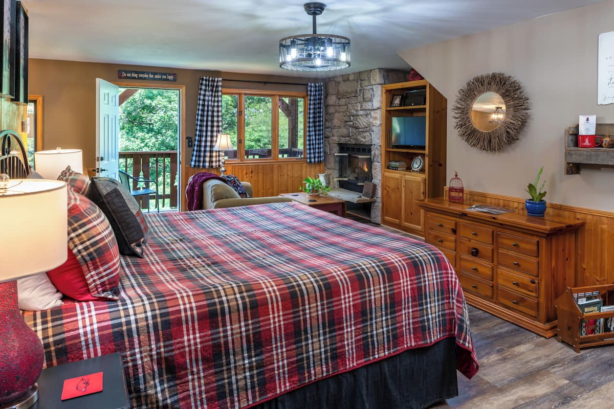 A cozy bedroom with a plaid bedspread, a fireplace, a wooden dresser, and a view of the outdoors.