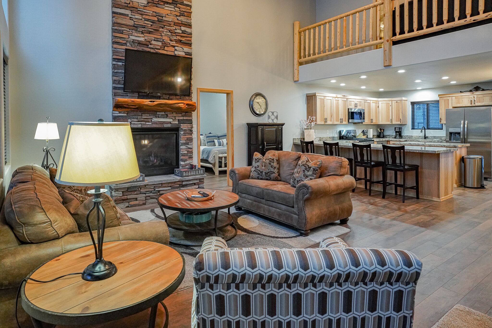 A spacious living area with a fireplace, a sofa, a coffee table, and a patterned ottoman. A kitchen with wooden cabinets and a stainless steel refrigerator is visible in the background.