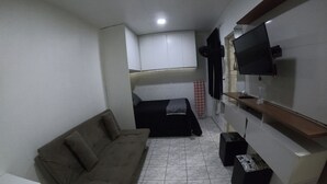Un espace de vie compact comprenant un canapé, un téléviseur fixé au mur et un petit coin cuisine.