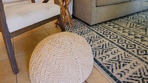 A living room with a grey sofa, a white armchair, a round woven ottoman, a small wooden side table with a plant, and a patterned rug. - Alpha @Serendip Vacation Condos (Cruz Bay)