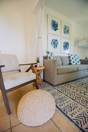 A living room with a grey sofa, a white armchair, a round woven ottoman, a small wooden side table with a plant, and a patterned rug. - Alpha @Serendip Vacation Condos (Cruz Bay)