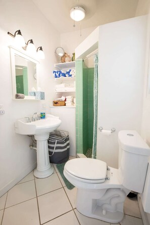 Een kleine badkamer met een wastafel op een voetstuk, toilet, douchegordijn en een spiegel met verlichting.