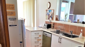A modern kitchen with a dishwasher, stove, and a small sink. There is a wooden floor and a window with blinds.
