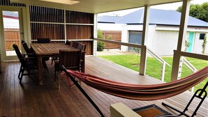 A covered patio with a wooden table and chairs, a hammock, and a view of a backyard with a lawn and a house.