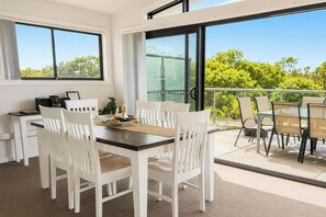 A dining area with a table and chairs, a coffee machine, and a view of greenery through large windows. - Beachside Beauty - Short walk to Caves Beach (Caves Beach)
