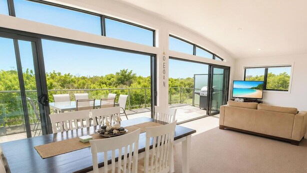 A modern living room with a dining area, large windows, a TV, and a balcony with a view. - Beachside Beauty - Short walk to Caves Beach (Caves Beach)