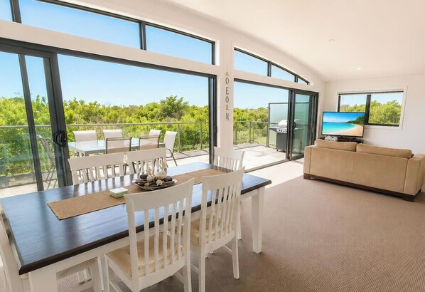 A modern living room with a dining area, large windows, and a view of greenery. - Beachside Beauty - Short walk to Caves Beach (Caves Beach)