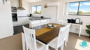 A modern kitchen with white cabinetry, a central island with a sink, and a dining area with a wooden table and white chairs. - Beachside Beauty - Short walk to Caves Beach (Caves Beach)