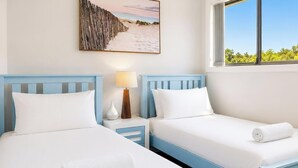 A hotel room with two beds, a nightstand with a lamp, a beach-themed picture on the wall, and a window with a view of greenery. - Beachside Beauty - Short walk to Caves Beach (Caves Beach)