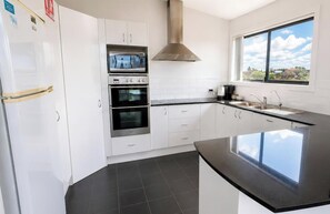 A modern kitchen with white cabinets, a black countertop, a built-in oven, and a window with a view. - Beachside Beauty - Short walk to Caves Beach (Caves Beach)