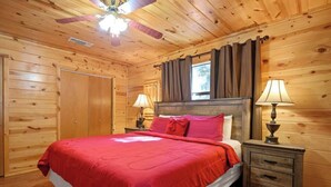 Camera da letto in una baita di legno, con un letto, due comodini con lampade, una ventola a soffitto e una finestra con tende.