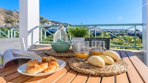 A table set with croissants, bread, a bowl, glasses of orange juice, and a book on a wooden table with a view of the sea and hills.