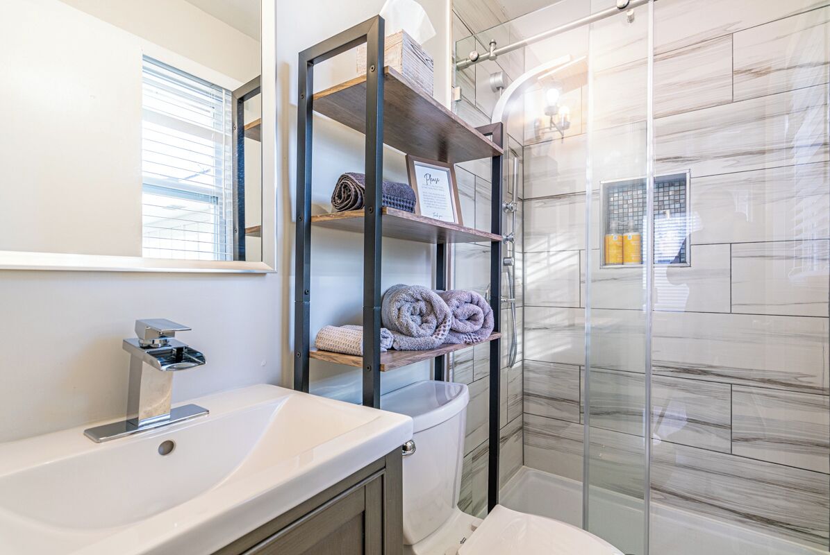 Ein modernes Badezimmer mit einem weißen Waschbecken, einer Duschkabine aus Glas und einem Holzregal mit Handtüchern.