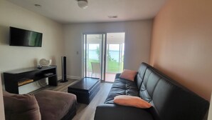 Un salon avec un canapé en cuir noir, un fauteuil marron, un téléviseur à écran plat, une grande enceinte sur pied et une porte-fenêtre coulissante donnant sur un balcon avec vue sur une étendue d’eau.