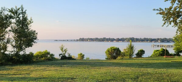 A lakeside view with a grassy area, trees, and a dock extending into the water. - Bright and airy waterfront getaway (Benedict)
