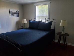 Une chambre à coucher avec un lit en métal, une couvre-lit bleue, deux tables de chevet avec des lampes, une fenêtre avec des stores et un tableau encadré au mur.