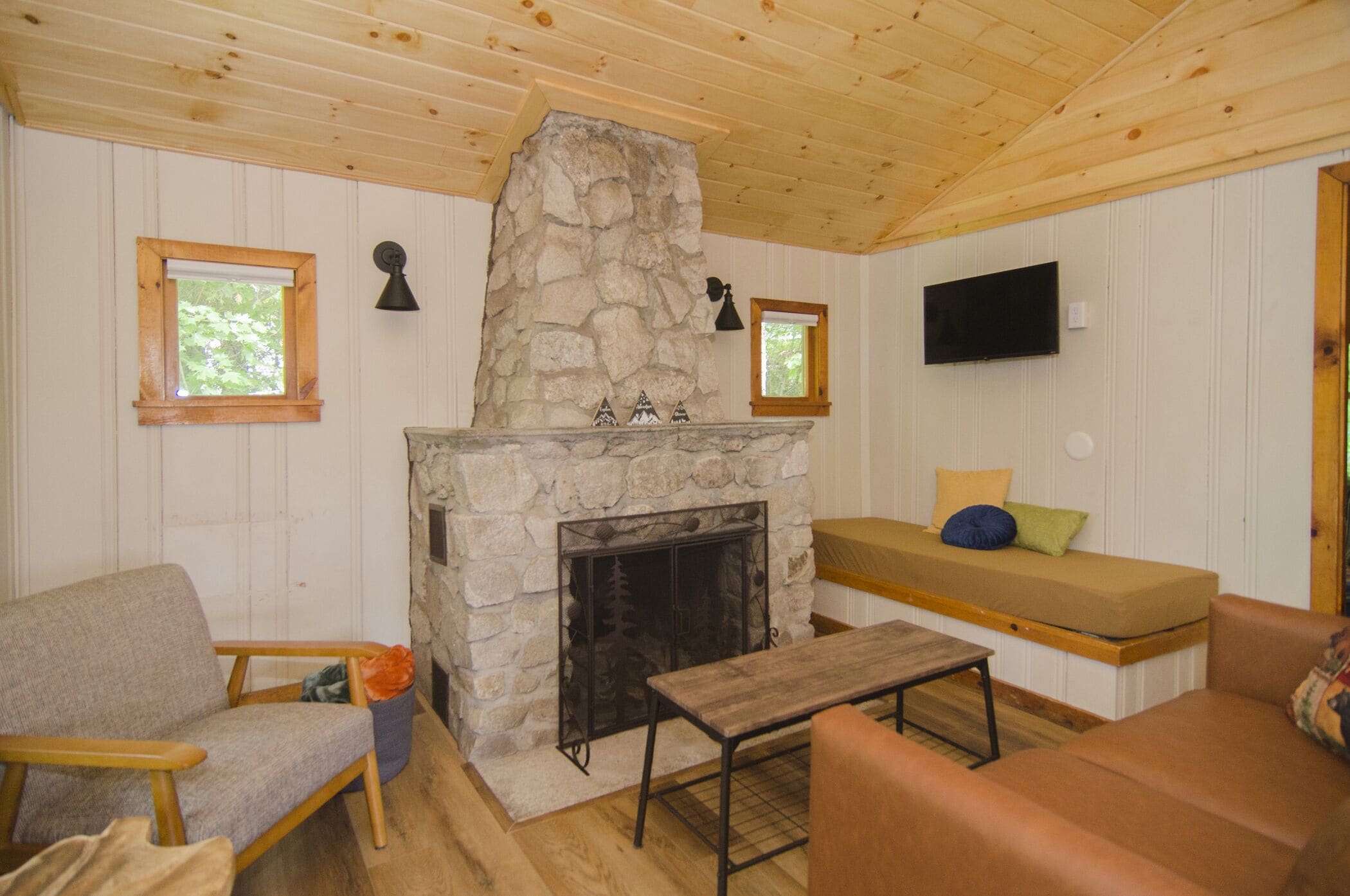 A cozy cabin interior with a stone fireplace, wooden furniture, and a television mounted on the wall.