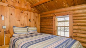 Un dormitorio tipo cabaña de troncos con cama, ropa de cama a rayas, una ventana y paredes de madera.