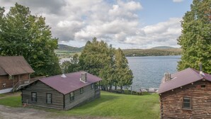 Una cabaña rústica con techo de metal, ubicada junto a un lago con un bosque al fondo.