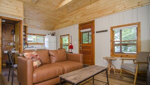 Un interior de cabaña acogedor, con techo de madera, un sofá de cuero marrón, una mesita de centro de madera y una zona de comedor con una lámpara roja.