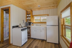A compact kitchen with wooden walls and a window, featuring a stove, refrigerator, and white cabinets. - Mountain Lake Cottages (Orleans)