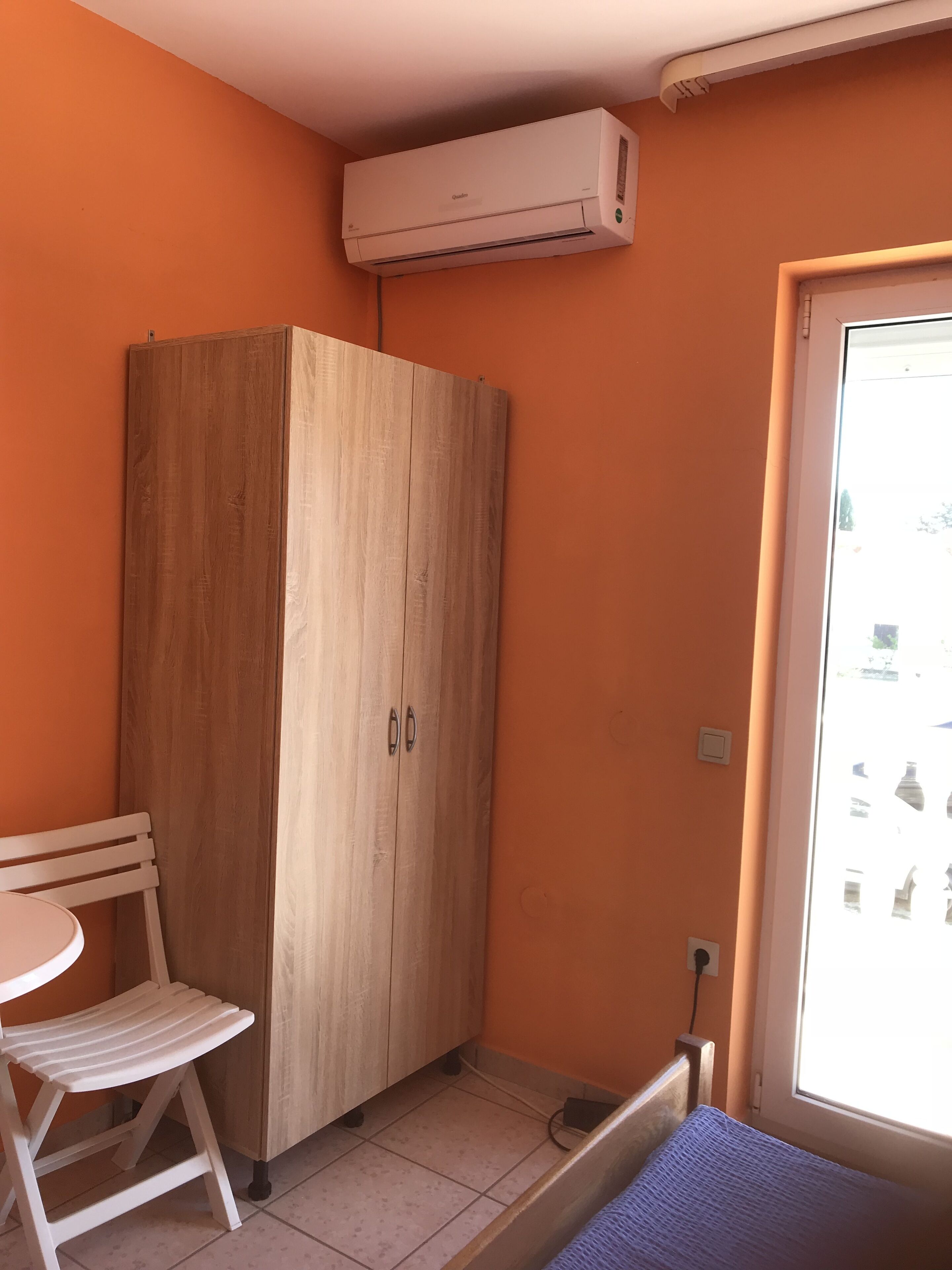 A small room with an orange wall, a wooden wardrobe, a white chair, a small table, and a window with a view.