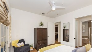 Une chambre à coucher avec un lit, un fauteuil sombre, une armoire, un ventilateur de plafond et une porte de salle de bain.