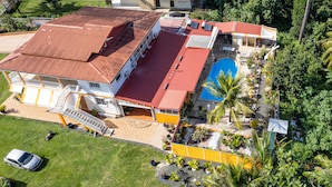 Vista aérea de um edifício de dois andares com telhado vermelho, piscina e um jardim bem cuidado com palmeiras.