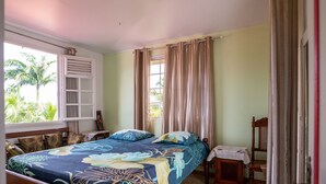Quarto com cama, mesa de cabeceira, cadeira, janela com cortinas e vista para palmeiras lá fora.