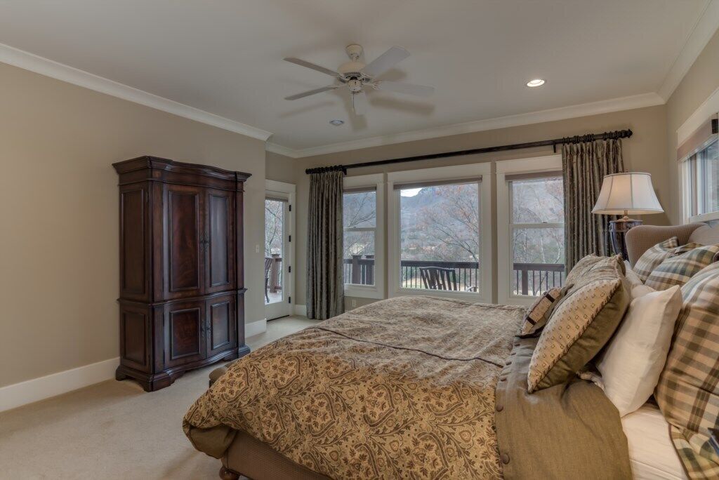 A bedroom with a large bed, a ceiling fan, a dark wooden cabinet, and a view of the outdoors through large windows.