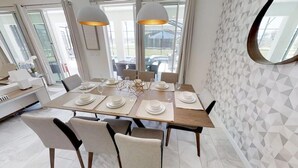 A modern dining area with a wooden table set for six, surrounded by chairs. There are pendant lights above the table, and the room has large windows offering a view of the outdoors.
