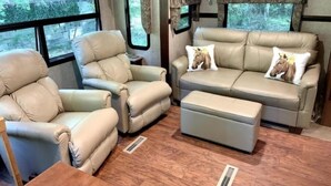 A cozy RV interior with leather seating, a sofa, and a window providing a view of greenery. - A Country Escape (Ayden)