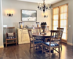 A dining area with a round table, six chairs, a chandelier, a painting, and a window with blinds.