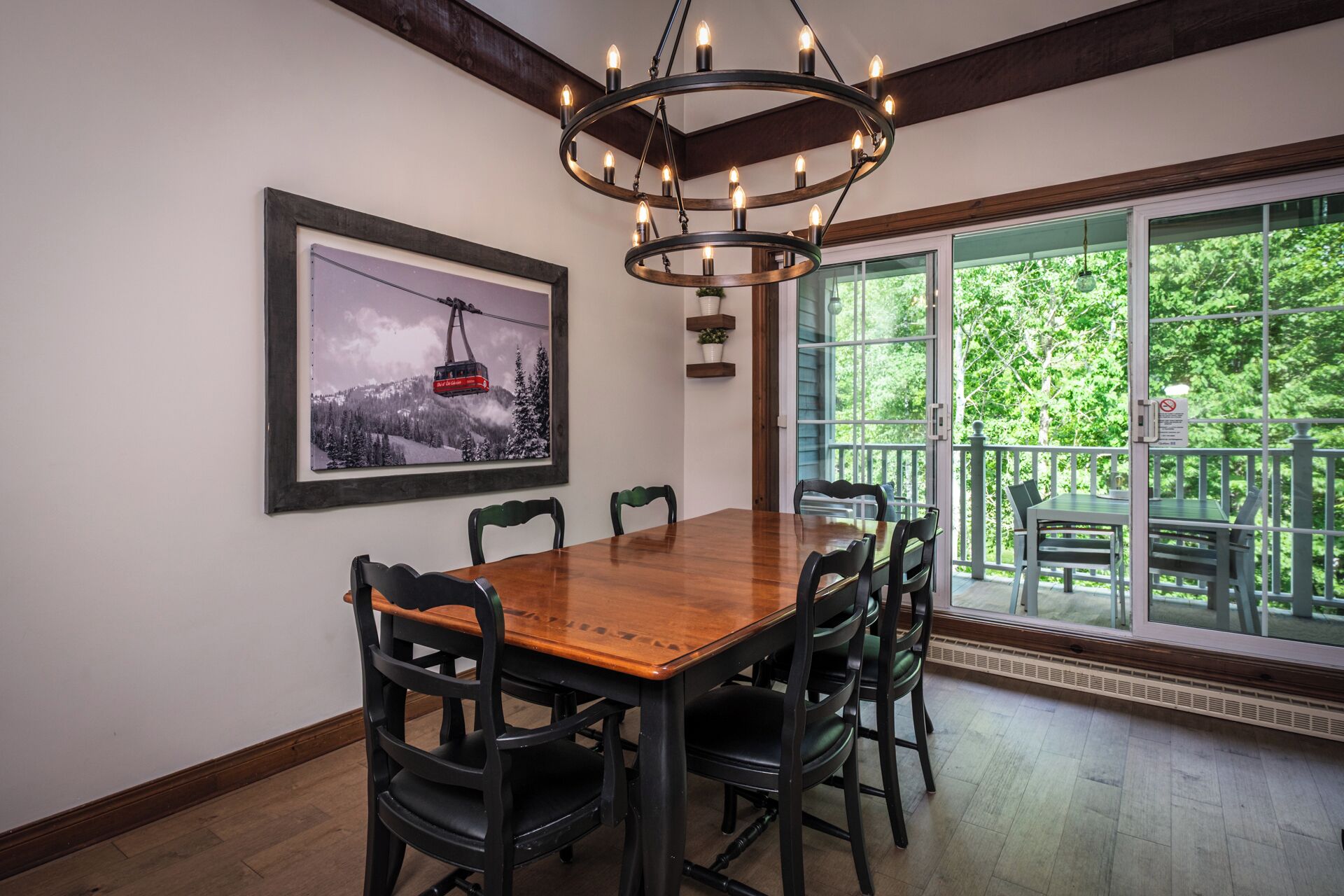 Un coin repas avec une table et des chaises en bois, un lustre, un tableau encadré représentant un télésiège et une vue sur la verdure à travers de grandes fenêtres.