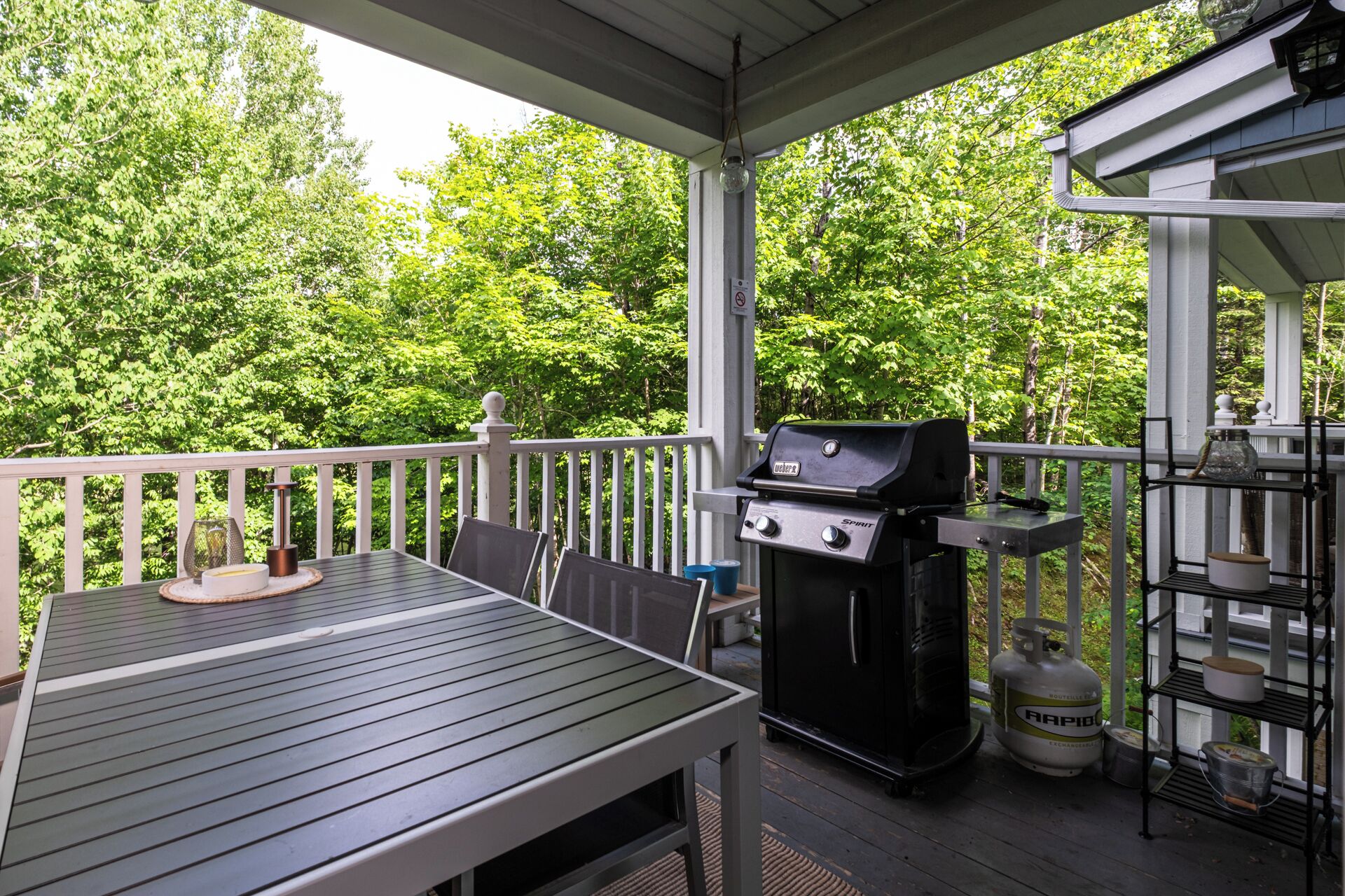 A covered outdoor deck with a grill, a table, chairs, and a gas cylinder.