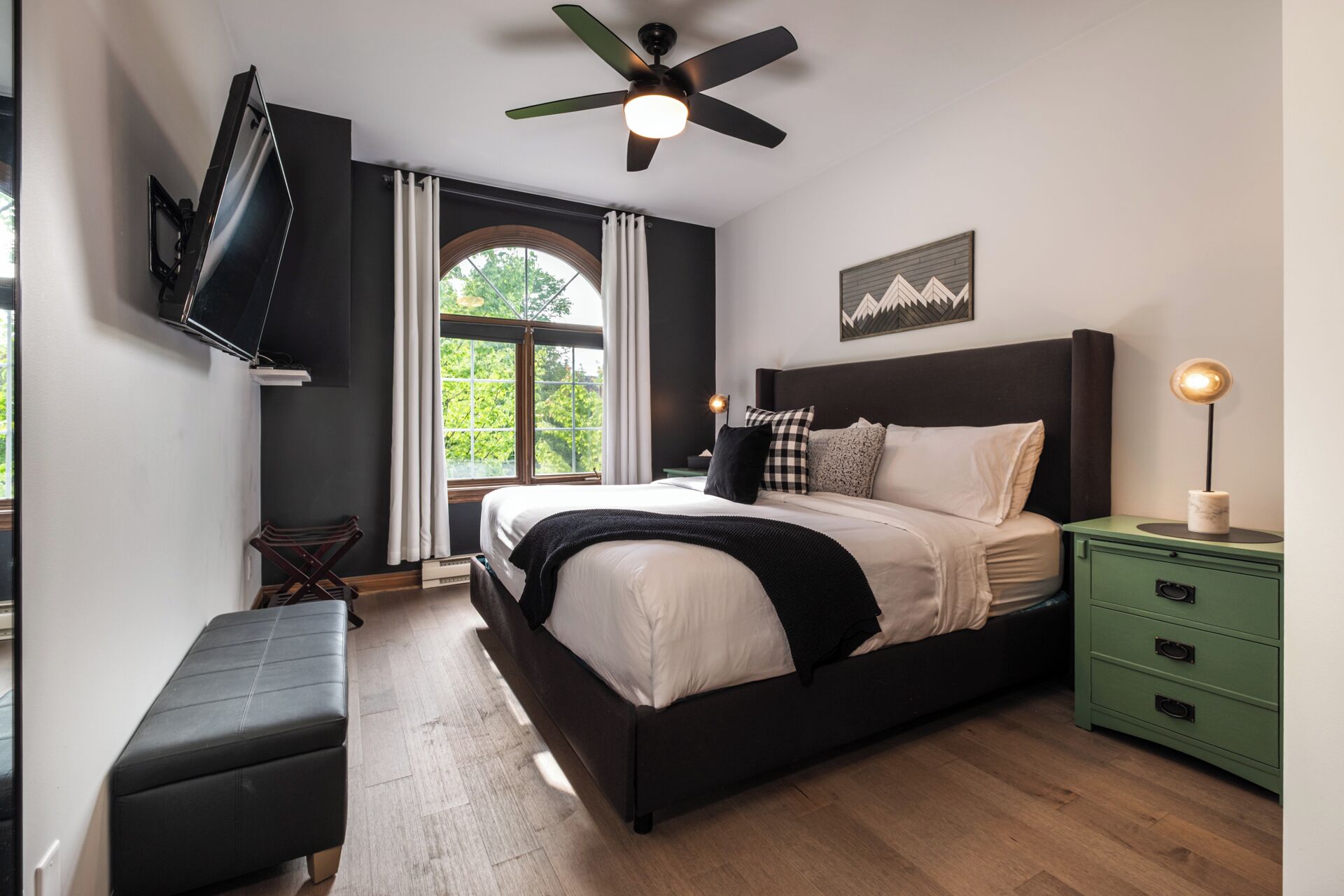 A modern bedroom with a large bed, a ceiling fan, a green nightstand, and a view of greenery outside.