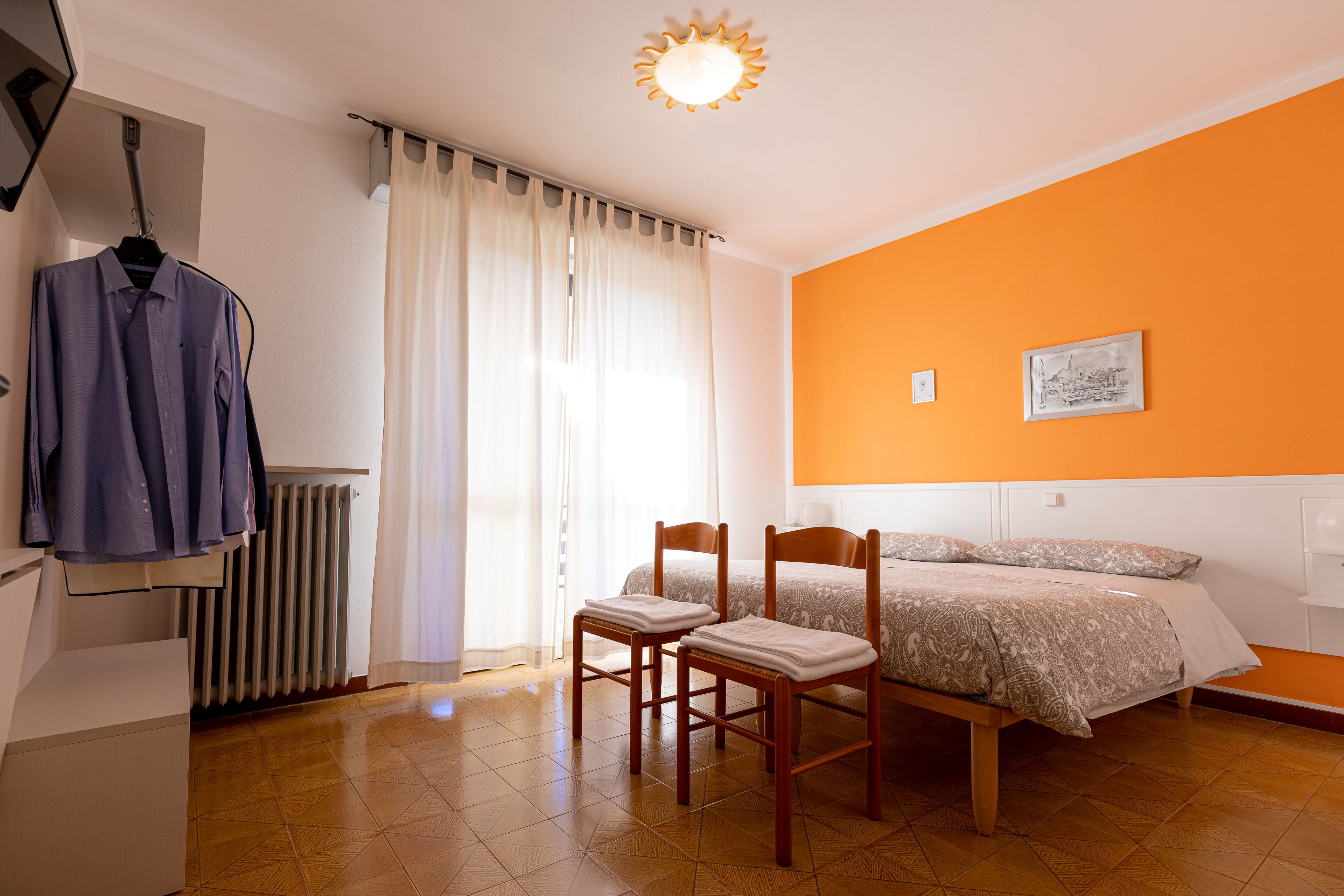 Un dormitorio con una cama, dos sillas, un radiador, una ventana con cortinas y una pared con un cuadro.