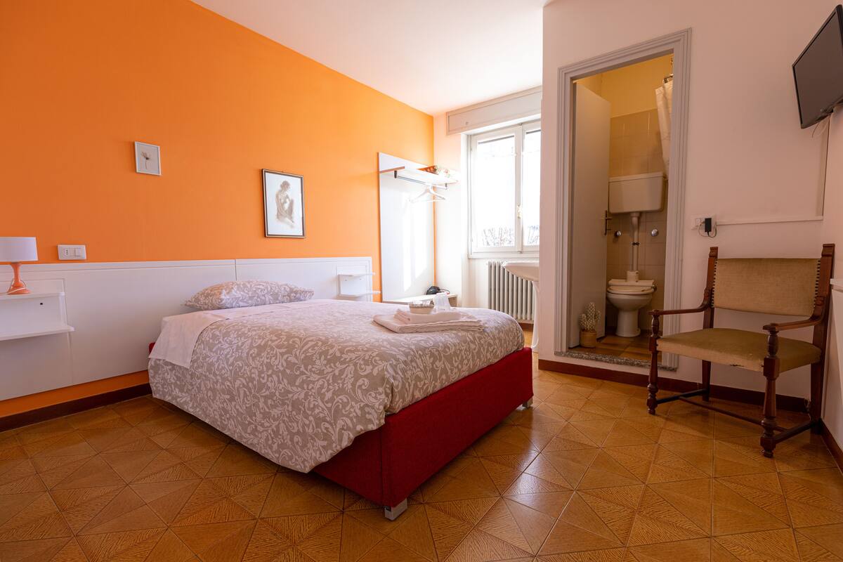 A bedroom with a red bed, a wooden chair, a white shelf, and a window with a view of the bathroom.