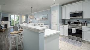 A modern kitchen with white cabinetry, a large island, stainless steel appliances, and a dining area with wicker stools.