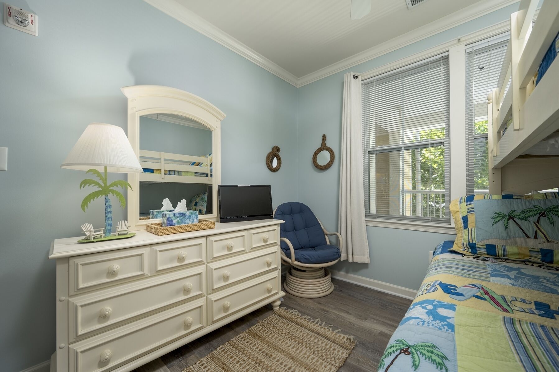 Un dormitorio con una cama litera, un escritorio con una computadora portátil, una silla, un aparador, una ventana con persianas y una colcha con motivos playeros.