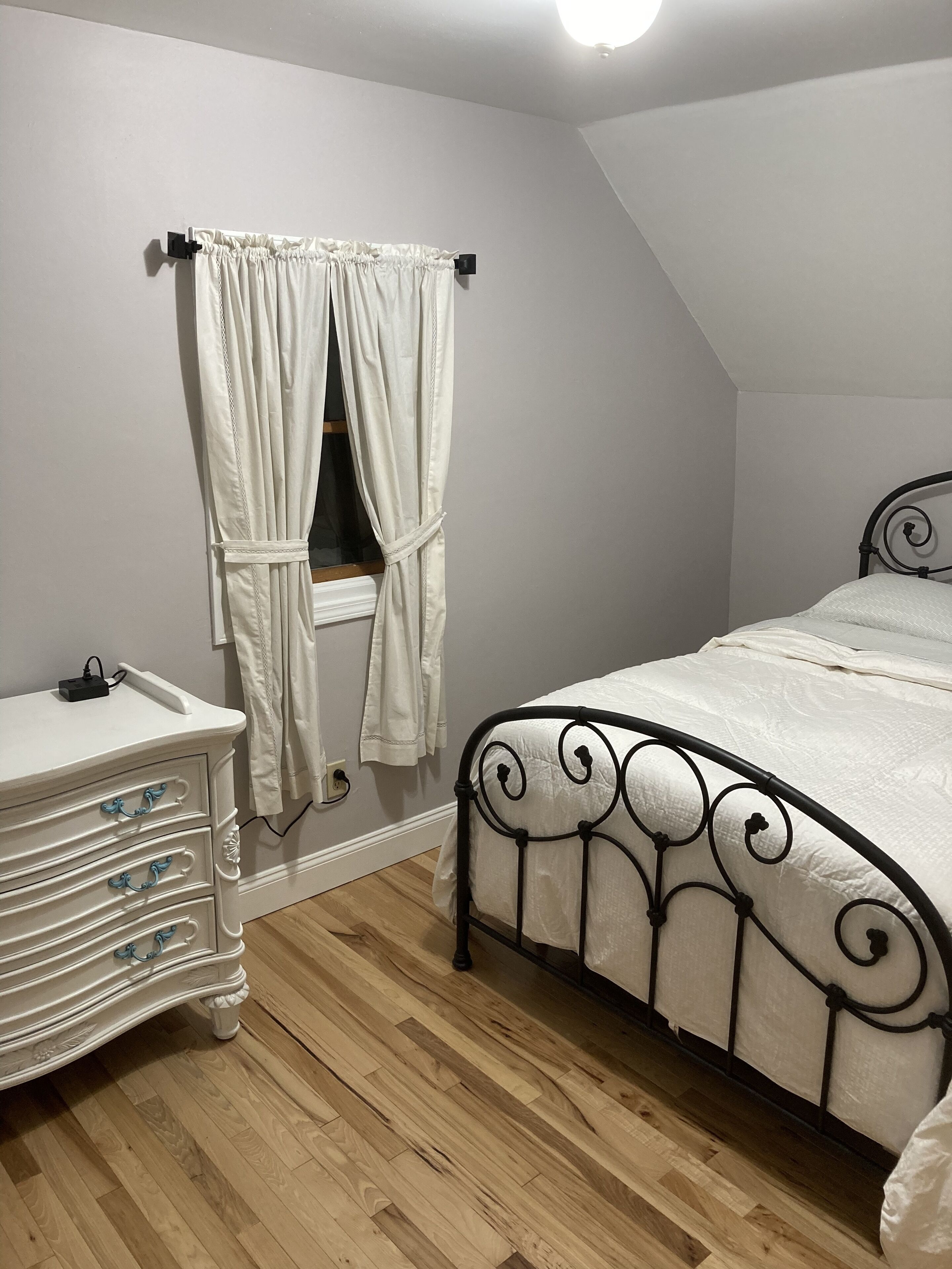 Un dormitorio con cama, cómoda, ventana con cortinas y lámpara en el techo.