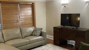 Uma sala de estar com sofá em formato de L, uma poltrona verde, uma televisão sobre um móvel de madeira e uma janela com persianas.