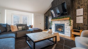 Una sala de estar moderna con chimenea, un televisor de pantalla plana grande montado en la pared y una zona de estar acogedora con un sofá seccional y otomanas.