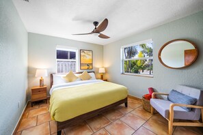 Un dormitorio con cama, mesitas de noche, ventilador de techo, ventana con vistas, espejo y una silla.