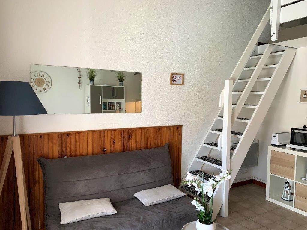 A cozy room with a grey sofa, a wooden headboard, a mirror, a staircase, and a small kitchen area.